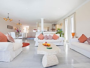 Ein geräumiges Wohnzimmer mit weißen Sofas, einem Couchtisch mit Früchten und einer modernen Küche im Hintergrund.