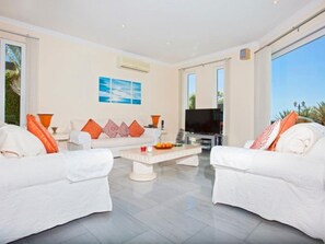 Ein modernes Wohnzimmer mit einer weißen Sofagarnitur, einem Couchtisch, einem Flachbildfernseher und einem großen Fenster.