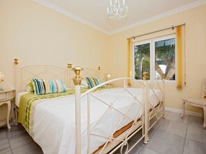Ein Bett mit weißem Metallgestell und goldenen Akzenten, ein Nachttisch mit Lampe sowie ein Fenster mit gelben Vorhängen.