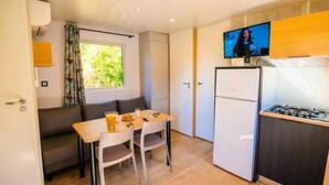 A compact kitchen with a refrigerator, a dining table with chairs, a TV mounted above the stove, and a window with curtains. - Cottage with AC - 6 guests (Vic-la-Gardiole)