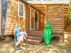 A wooden cabin with a porch, a person sitting on a chair, and a green alligator statue. - Cottage with air conditioning - 6 guests (Vic-la-Gardiole)