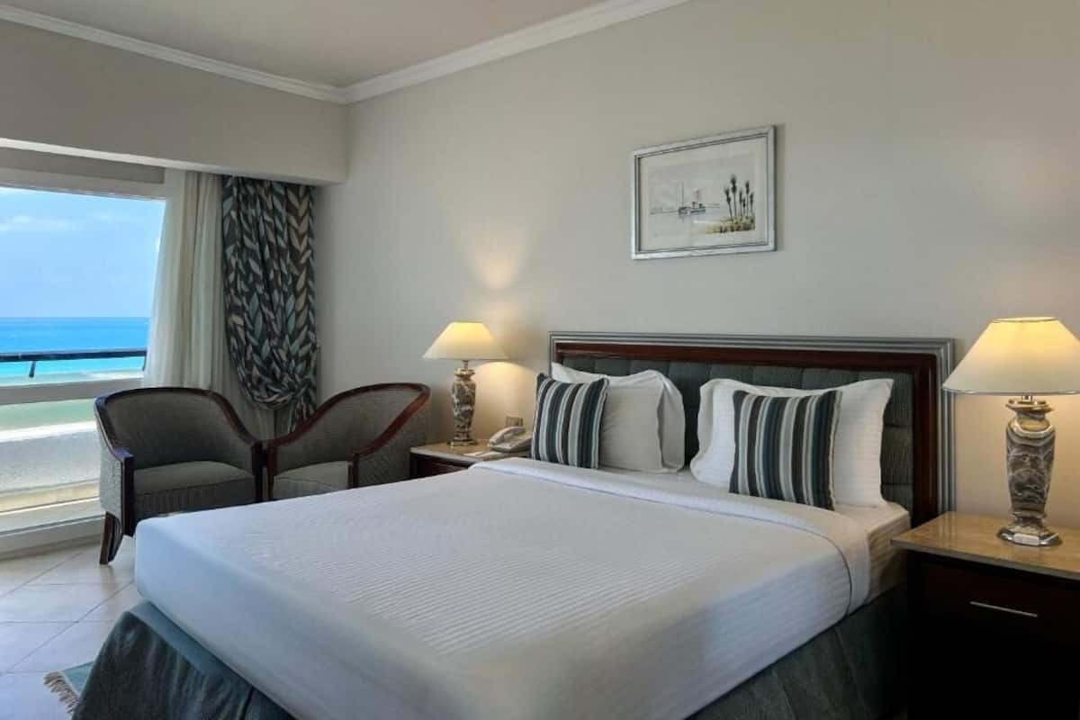 A hotel room with a bed, two bedside lamps, a chair, a nightstand, a framed picture on the wall, and a view of the ocean.