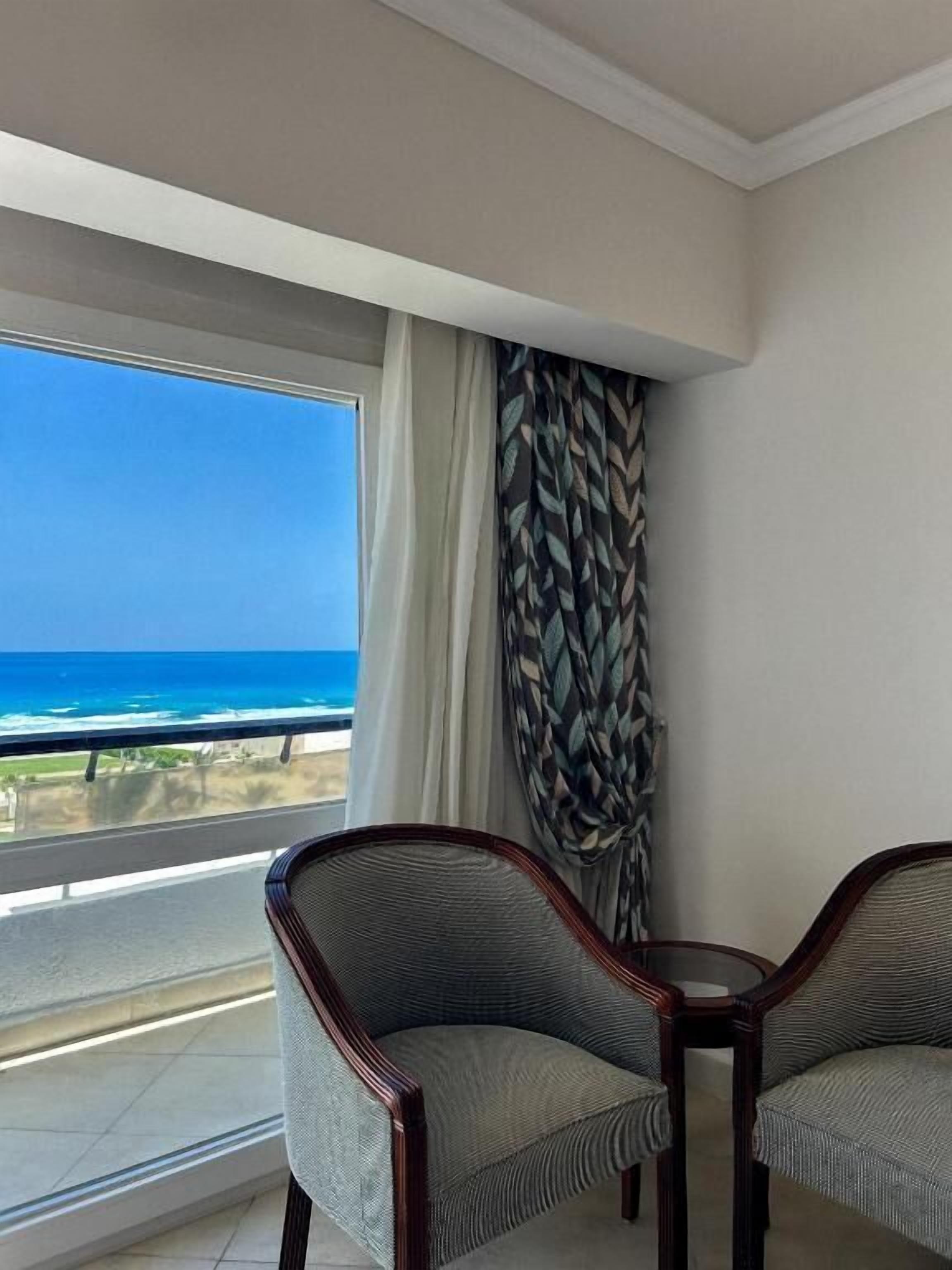 A room with a large window overlooking the ocean, two upholstered chairs, and a patterned curtain.