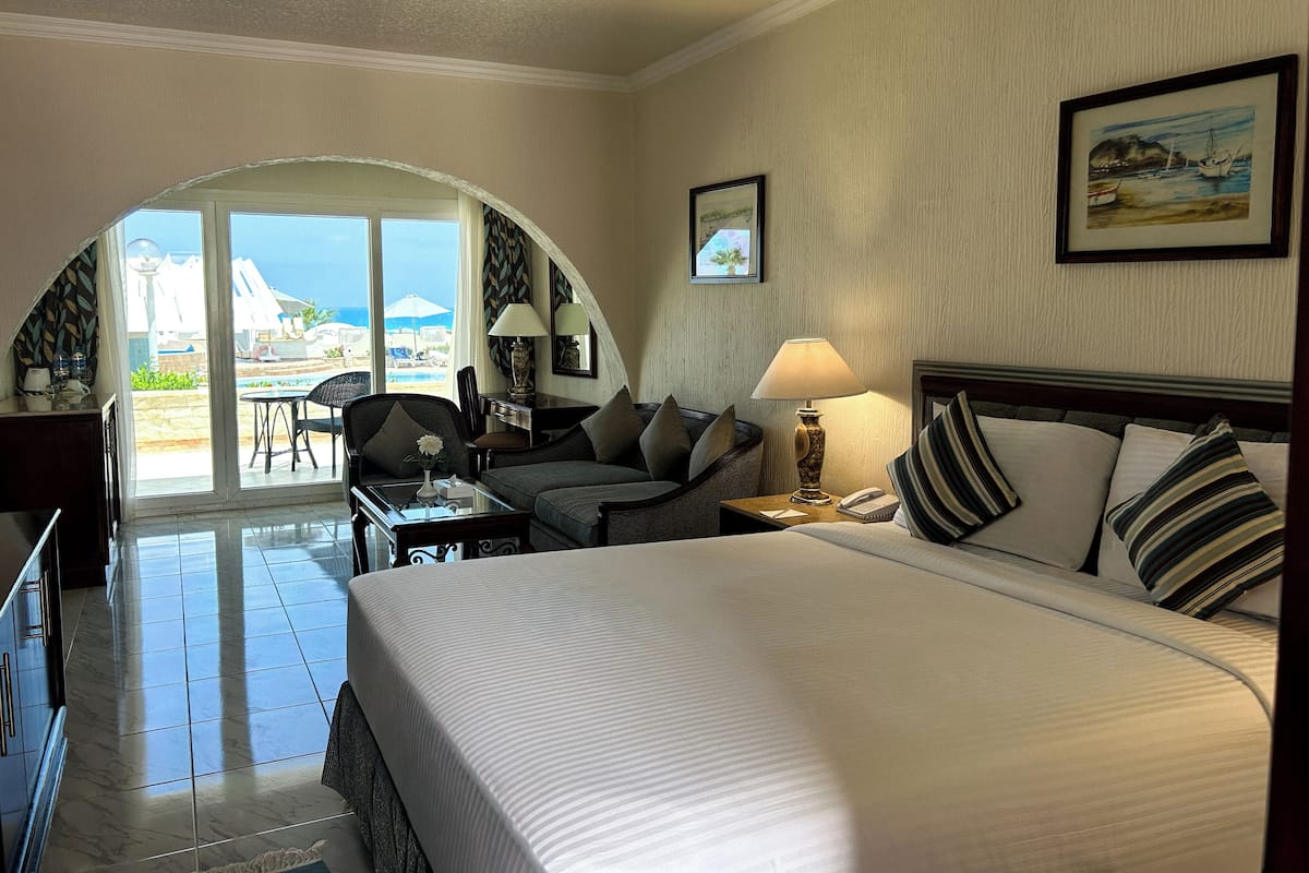 A hotel room with a bed, a desk, a chair, a lamp, and a view of the pool and beach.