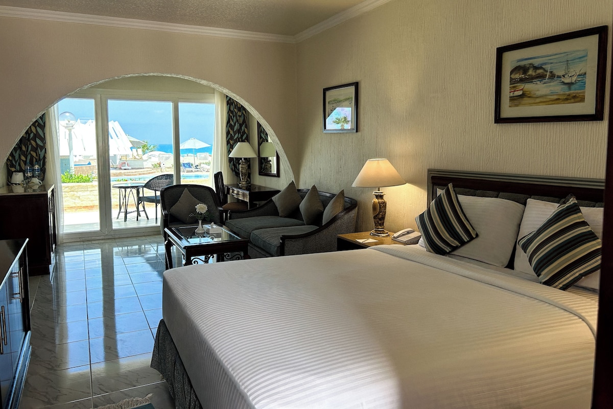 Habitación de hotel con cama, escritorio, silla, lámpara y vista a la piscina y la playa.