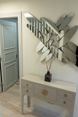 Ein Zimmer mit einer weißen Kommode, einem dekorativen Spiegel mit blattartigen Formen und einer kleinen Vase mit Schilfrohren.