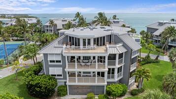 Um edifício residencial com vários andares e varanda, rodeado por palmeiras e uma quadra de tênis.