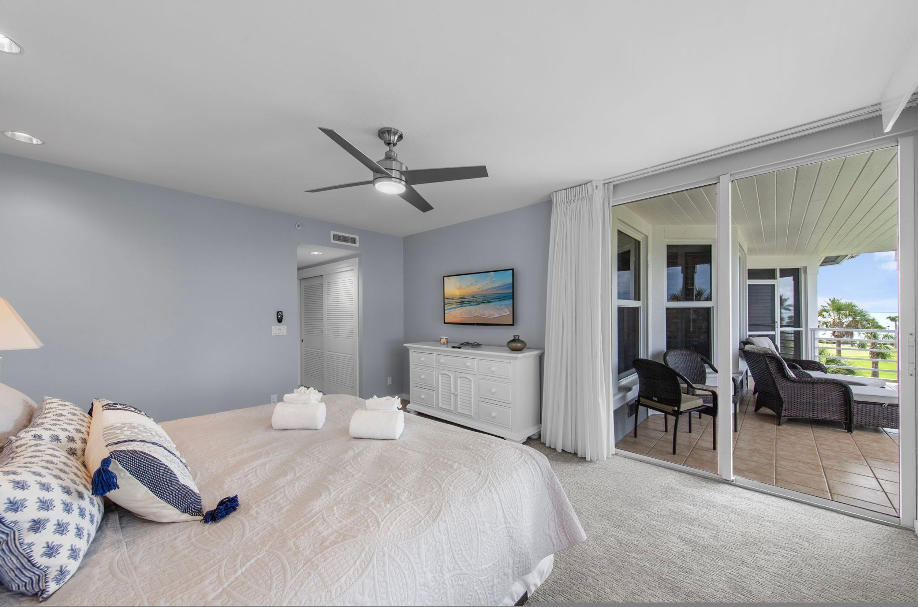 A bedroom with a bed, a ceiling fan, a nightstand, a dresser, a painting, and a sliding glass door leading to a balcony with outdoor furniture.