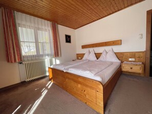 A bedroom with a wooden bed, a nightstand, a radiator, and a window with curtains. - Room Josefine - Comfort room with mountain view, Julius Appartements Wagrain (Hofmark)