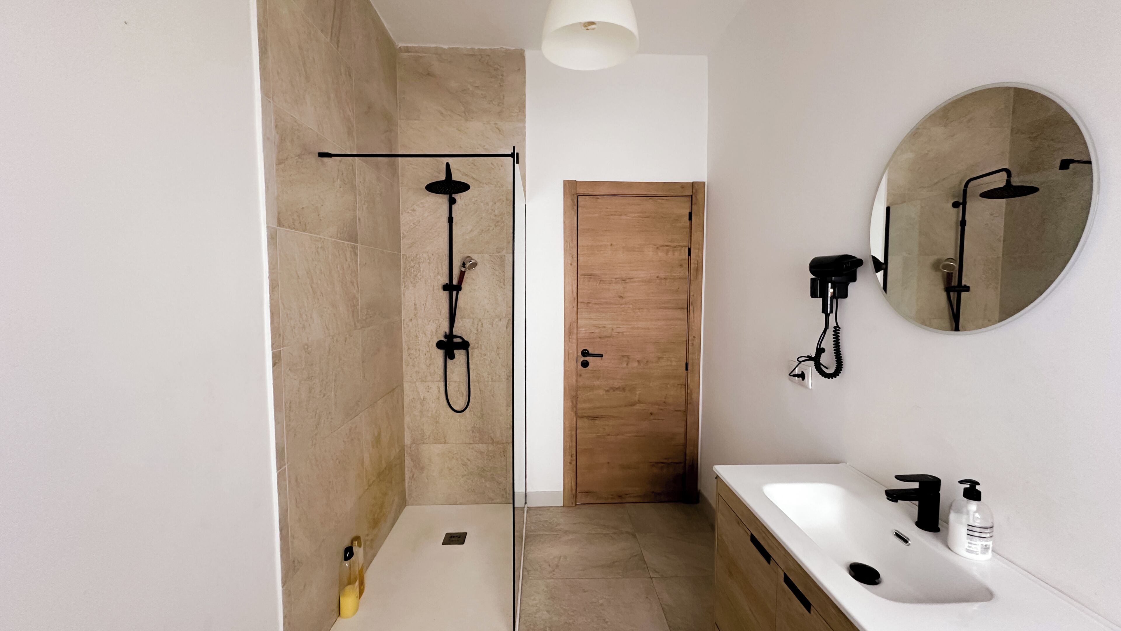A modern bathroom with a glass-enclosed shower, a wooden door, a white sink with a black faucet, and a round mirror.