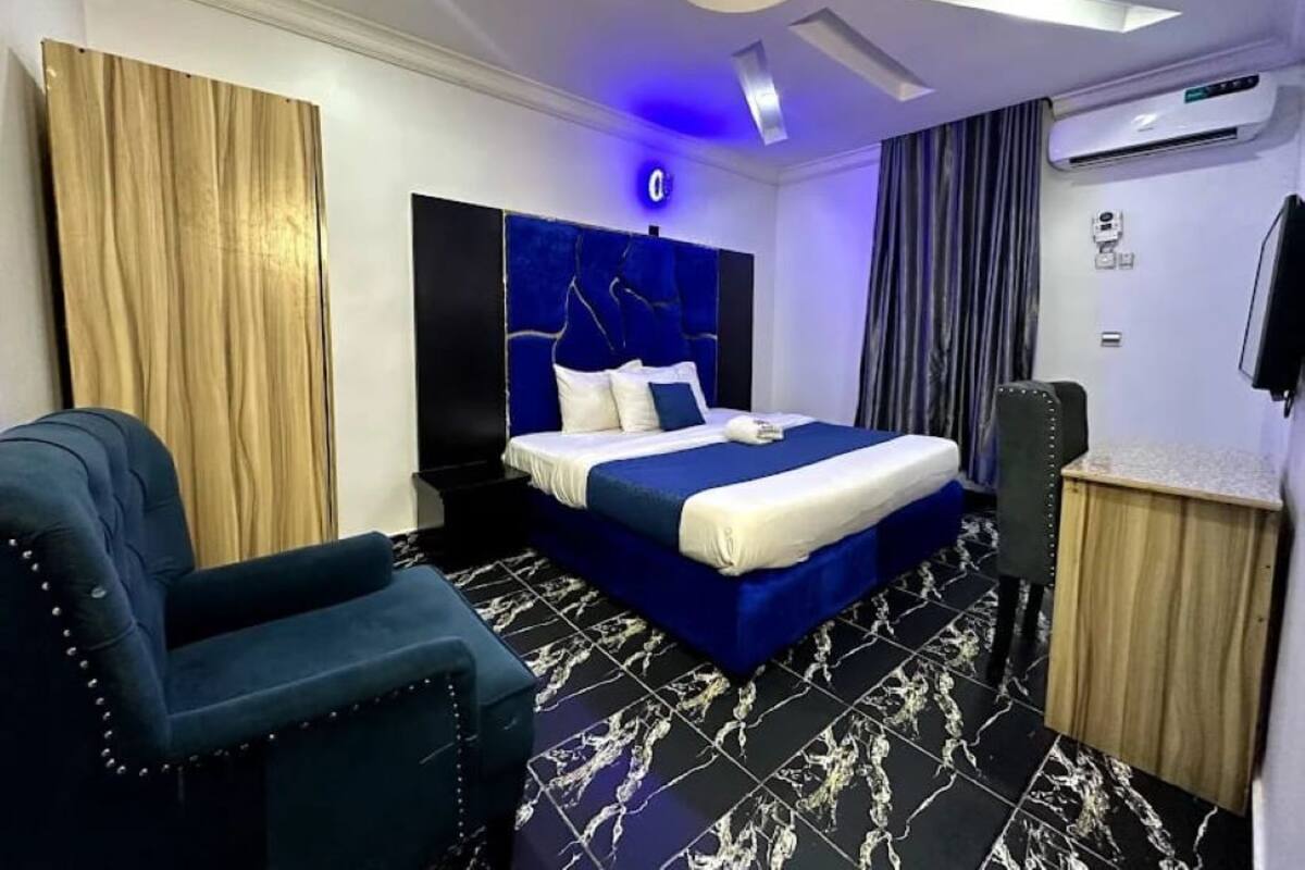 A modern hotel room with a bed, a blue armchair, a wooden cabinet, and a ceiling light fixture.
