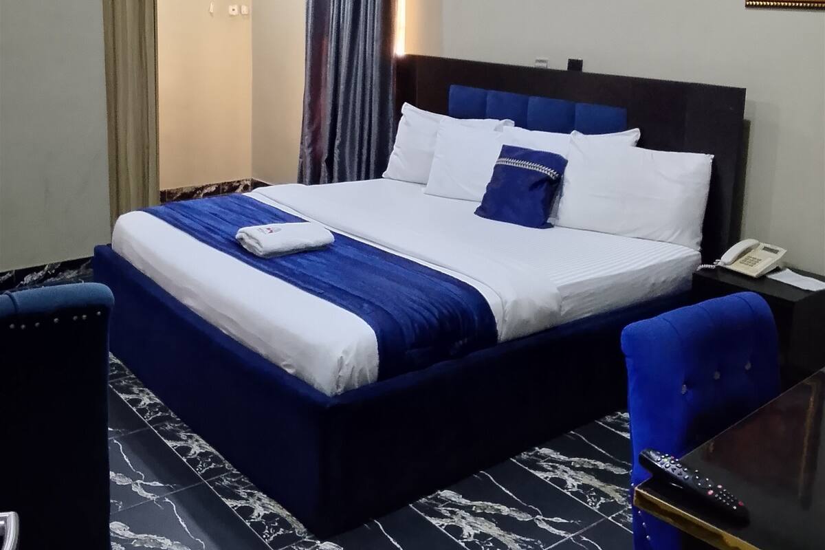 A hotel room with a bed, a desk, a chair, an air conditioning unit, a telephone, and a painting on the wall.