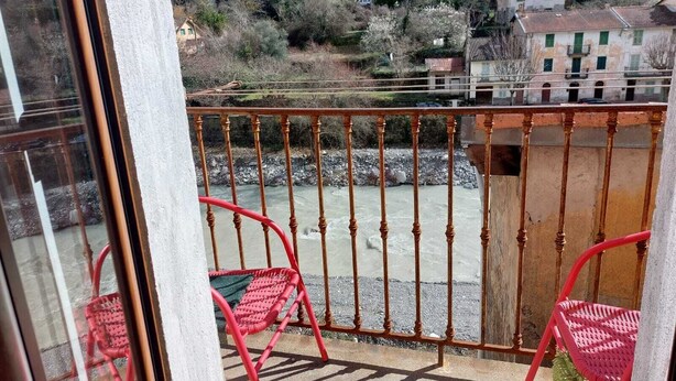 A balcony with a metal railing, two red chairs, and a view of a river and buildings in the background. - Furnished Appartment Au Pied du Mercantour (Fontan)