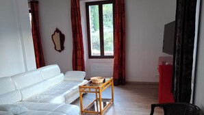 A living room with a white sofa, a wooden coffee table, a black chair, a television, and a window with curtains. - Furnished Appartment Au Pied du Mercantour (Fontan)