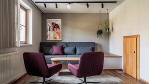 A modern living room with a sofa, two armchairs, a wooden coffee table, a radiator, a window with curtains, a hanging plant, and a door. - Room Tobias - Comfort room with mountain view, Julius Appartements Wagrain (Hofmark)