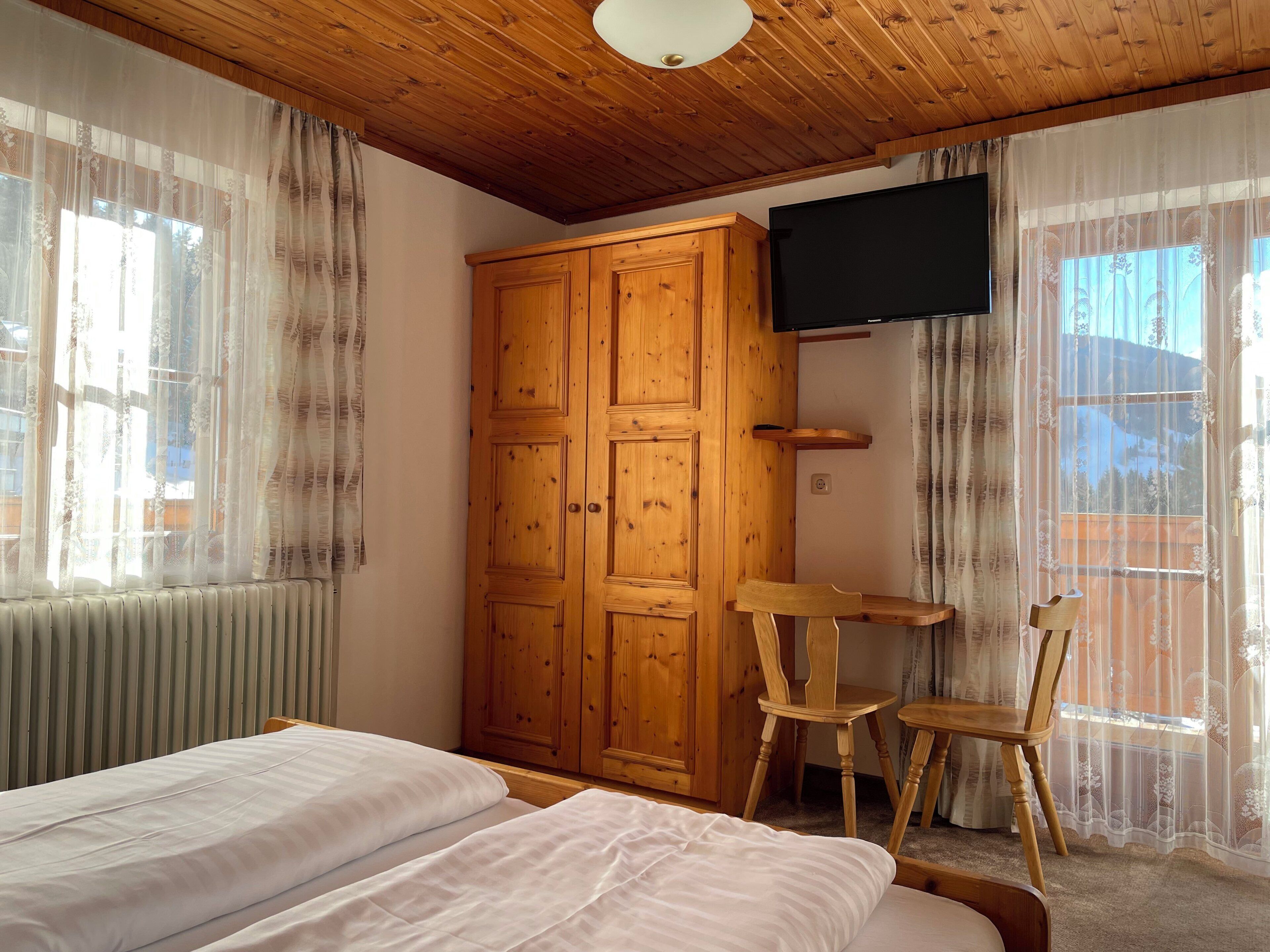 Een slaapkamer met een houten garderobe, een aan de muur bevestigde televisie, twee houten stoelen en een bed met wit beddengoed.