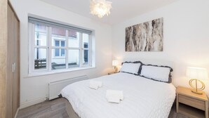 Un dormitorio con una cama grande, una ventana, un radiador, una mesita de noche y una obra de arte colgada en la pared.
