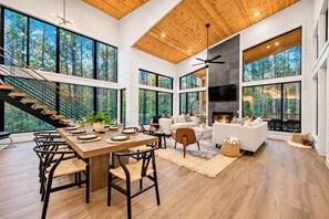 A spacious living area with a wooden floor, large windows, a fireplace, and a modern staircase. - Family Cabin | Hot Tub, Games & Fire | Sleeps 17 (Broken Bow)