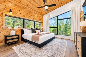 A bedroom with a large bed, a ceiling fan, and a view of trees through large windows. - Family Cabin | Hot Tub, Games & Fire | Sleeps 17 (Broken Bow)