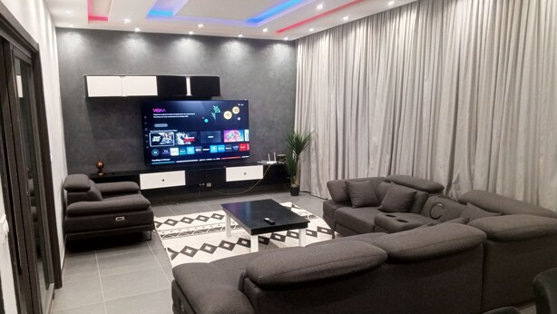 Ein modernes Wohnzimmer mit Fernseher, dunklen Sofas, einem Couchtisch und einem gemusterten Teppich.