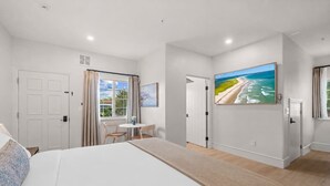 A hotel room with a bed, a small table with chairs, a painting of a beach, and a door leading to another room. - Bella Notte by the beach Studio 1 (Pleasure Point)