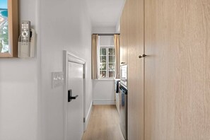 En smal hall med en trädörr, ett fönster med gardiner, en väggmonterad fjärrkontroll och ett köksområde med en spis och ugn.