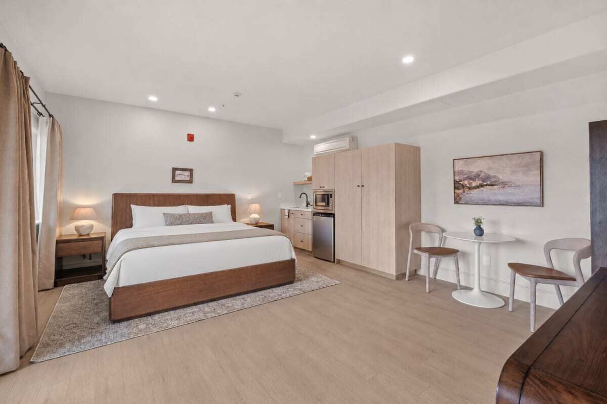 Moderni hotellihuone, jossa on puinen sänky, pieni keittiönurkkaus, pieni ruokailutila ja taulu seinällä.