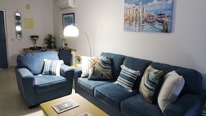 Een woonkamer met een blauwe bank, een houten salontafel, een plafondventilator en een schilderij van boten aan de muur.