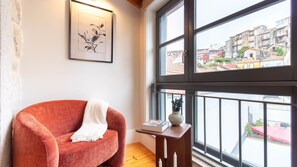 Ein Zimmer mit einem roten Sessel, einem gerahmten Kunstwerk, einem kleinen Tisch mit Buch und Vase sowie einem großen Fenster mit Blick auf Gebäude.