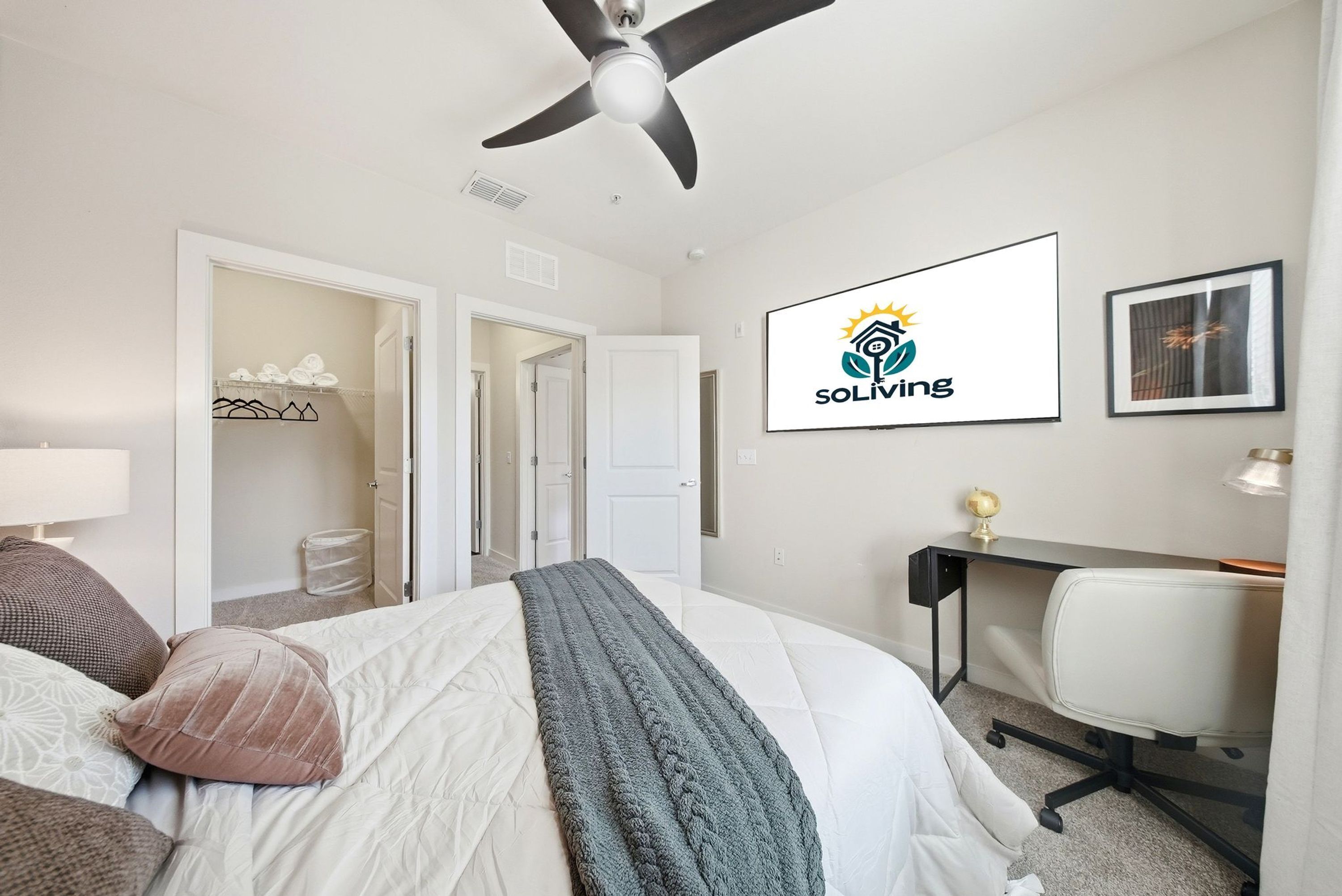 A hotel room with a bed, a desk, a chair, a ceiling fan, and a wall-mounted logo.