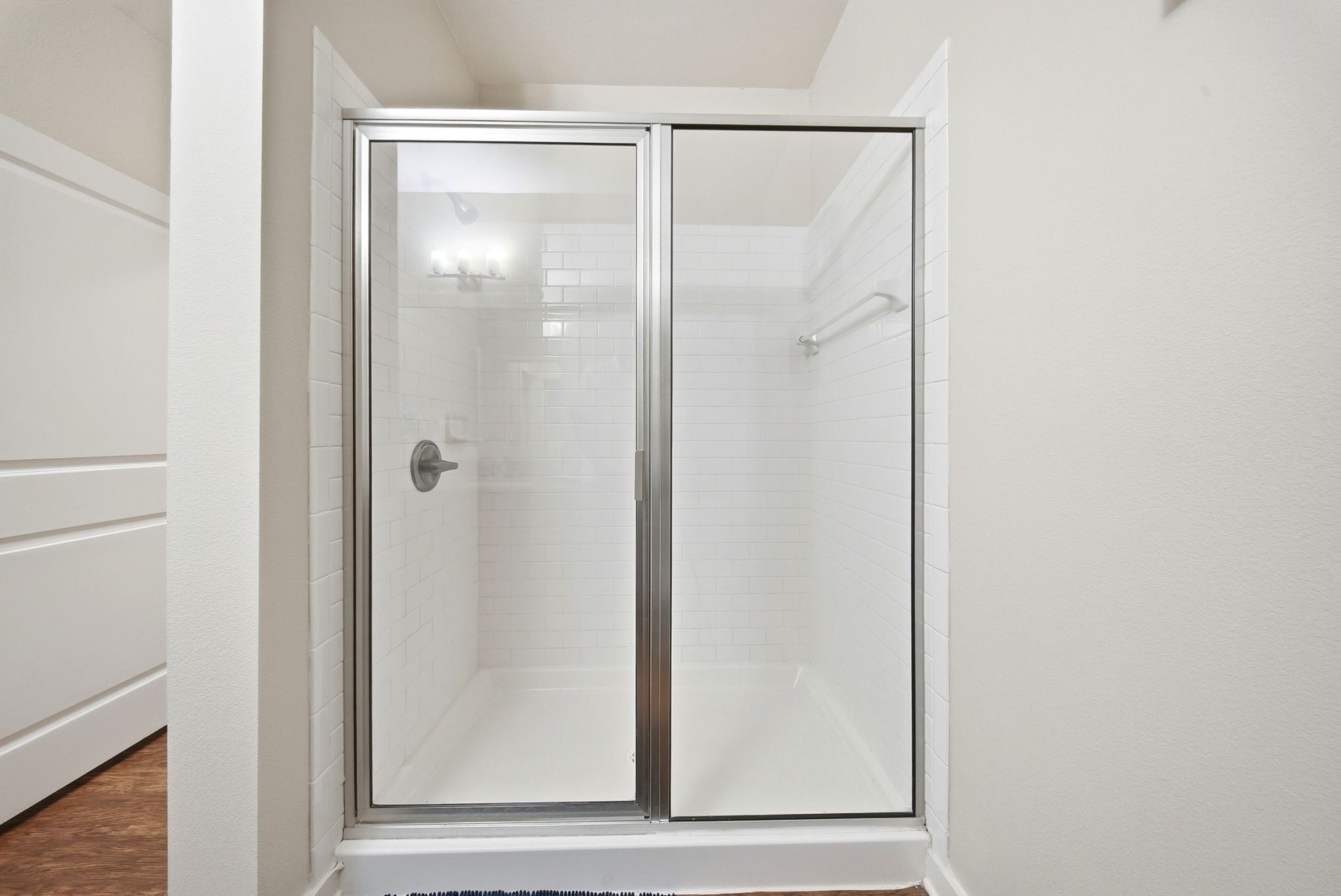Una ducha con cerramiento de vidrio, pared de azulejos blancos y puerta blanca.