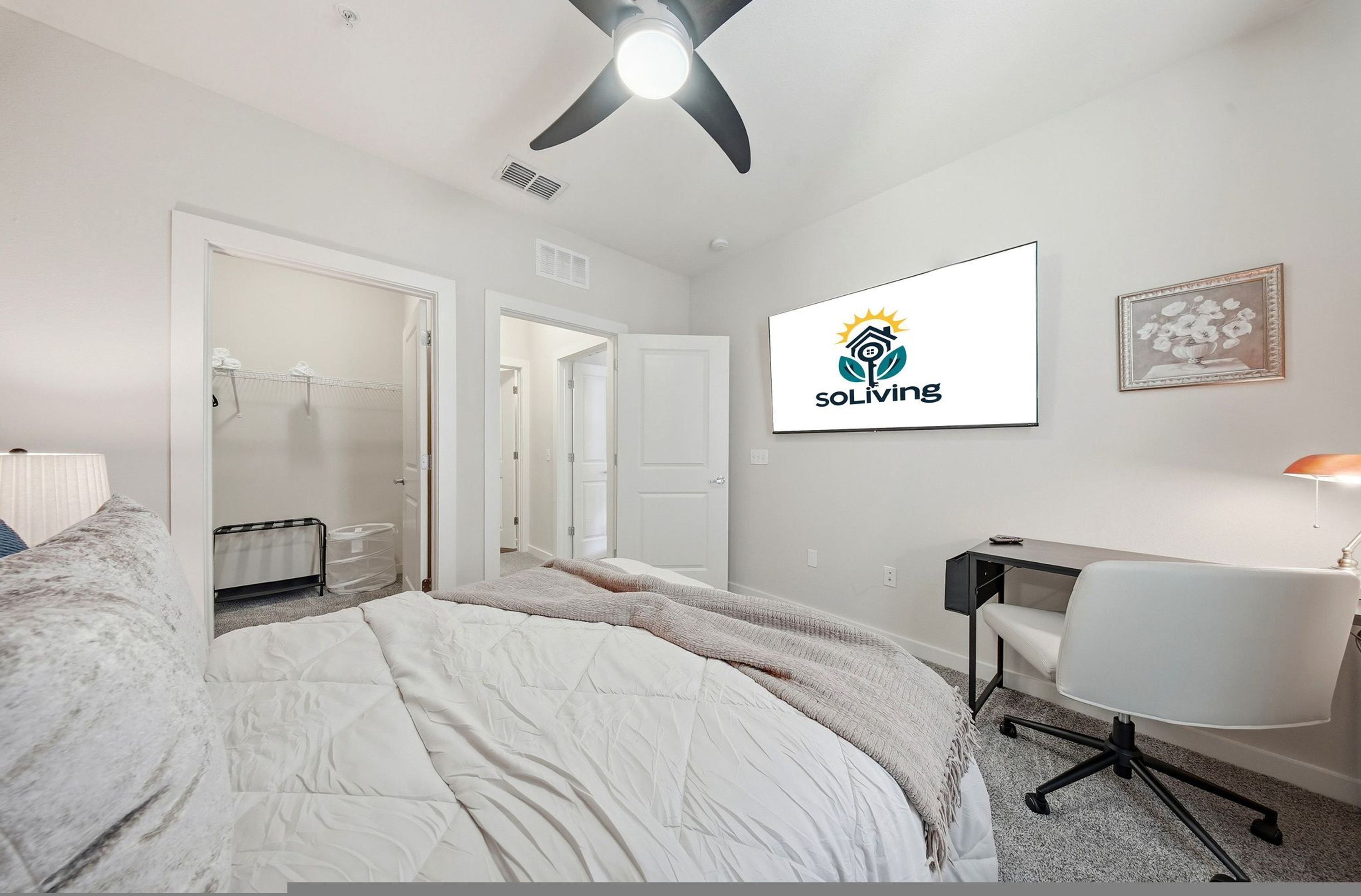 A bedroom with a bed, a desk, a chair, a ceiling fan, and a wall-mounted logo.
