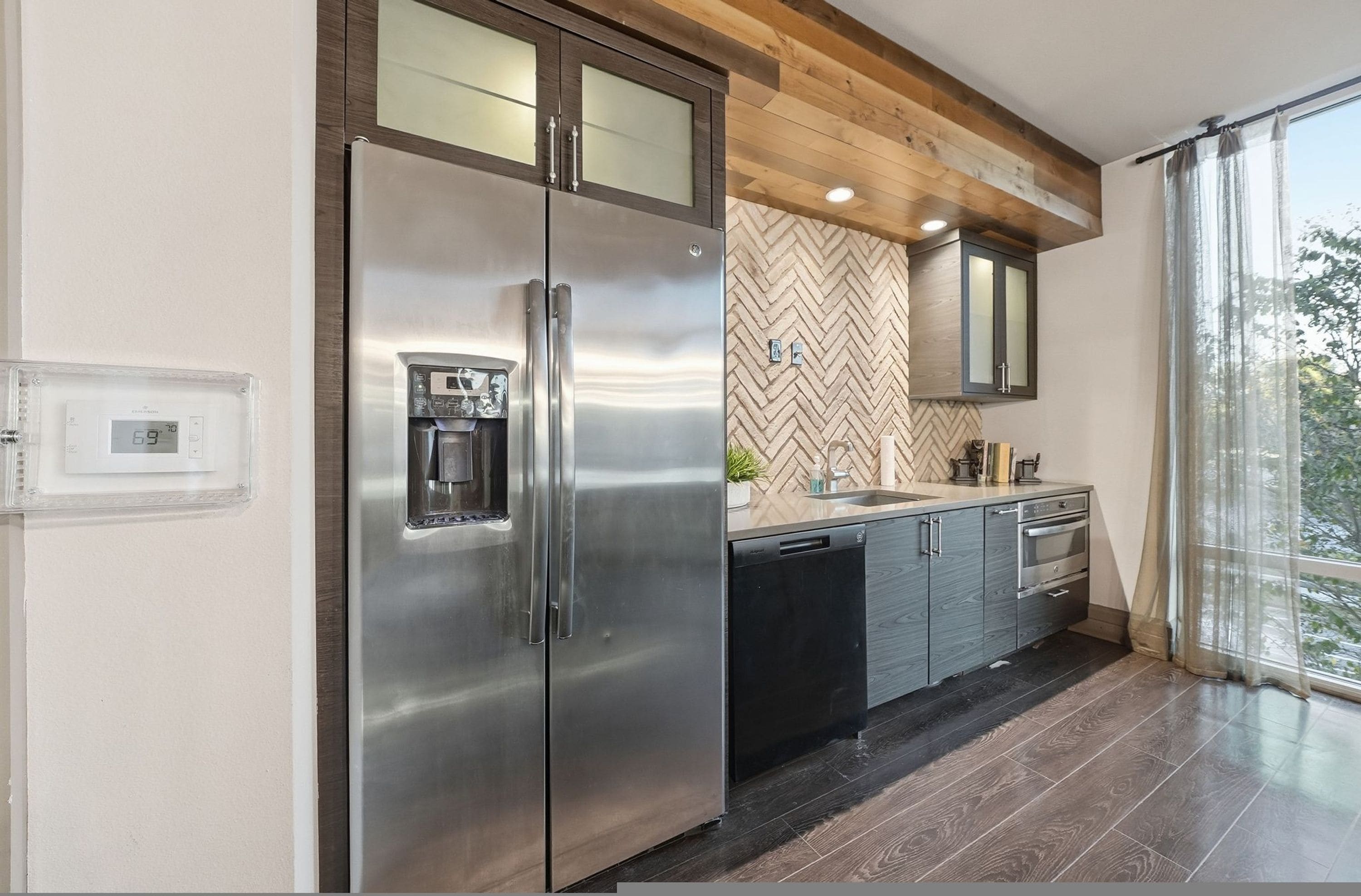A modern kitchen with a stainless steel refrigerator, wooden ceiling beams, a herringbone-patterned backsplash, and a large window with a view.