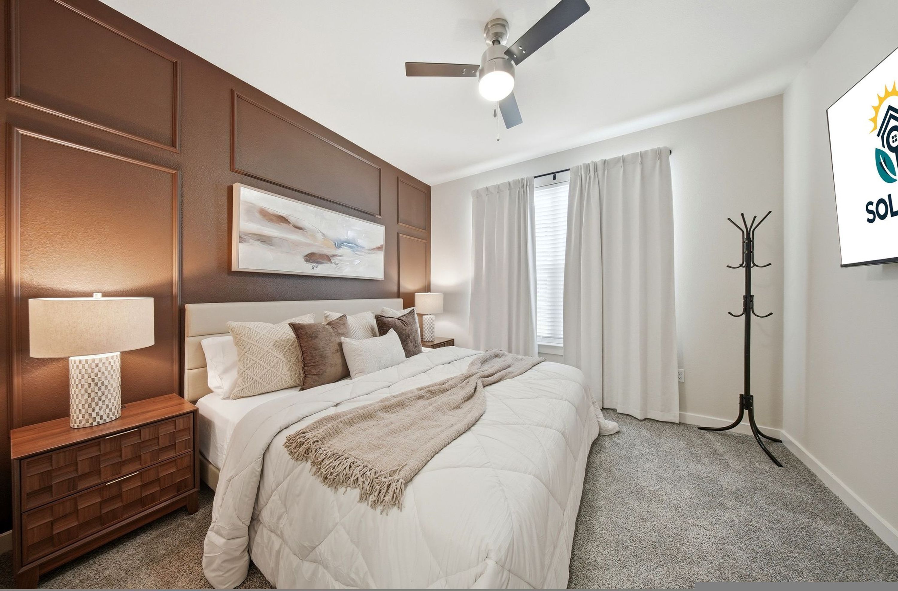 Une chambre à coucher comprenant un lit, des tables de chevet, un ventilateur de plafond, un porte-manteau et un tableau accroché au mur.