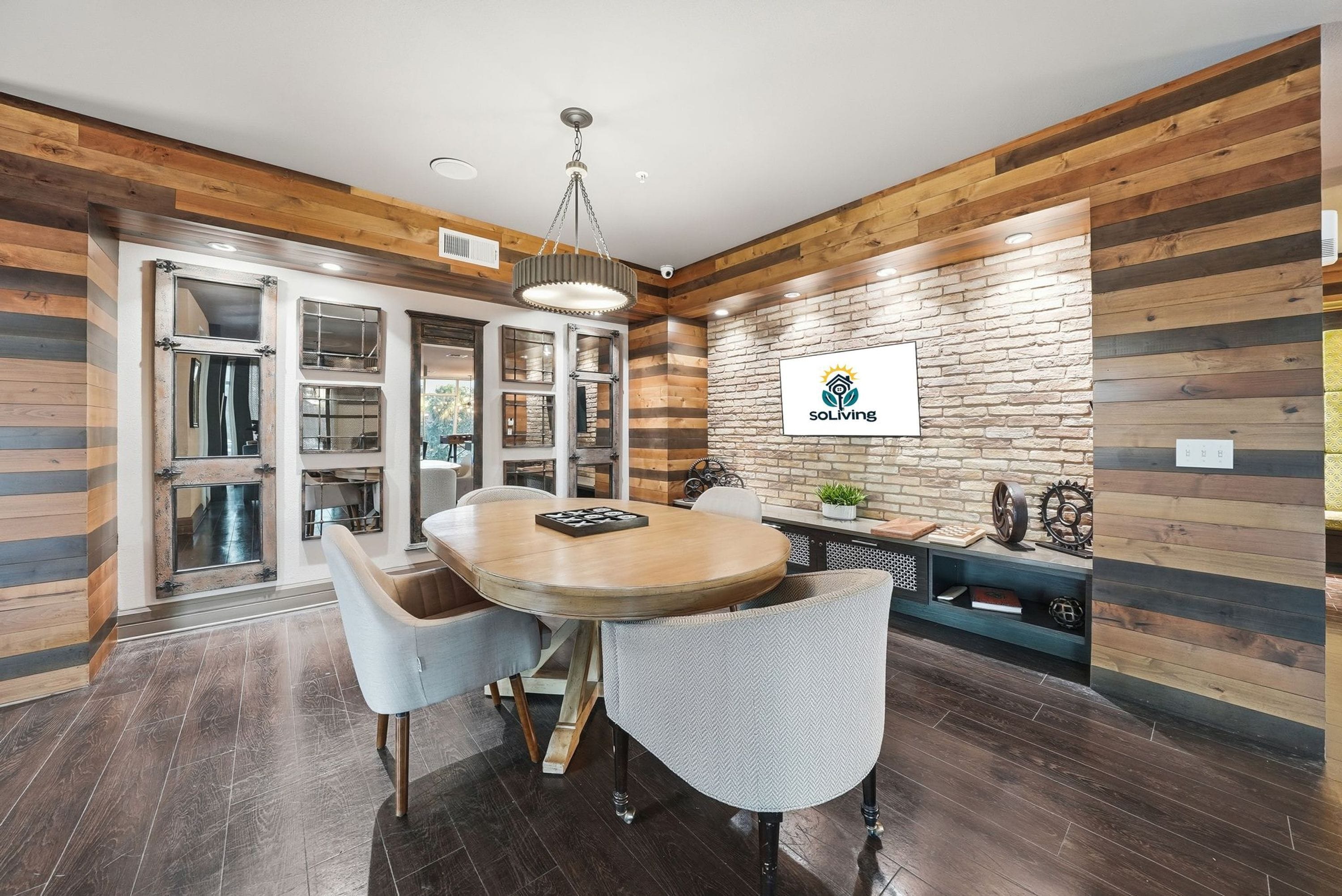 A modern dining area with a wooden table and chairs, a chandelier, and a brick wall with a logo.