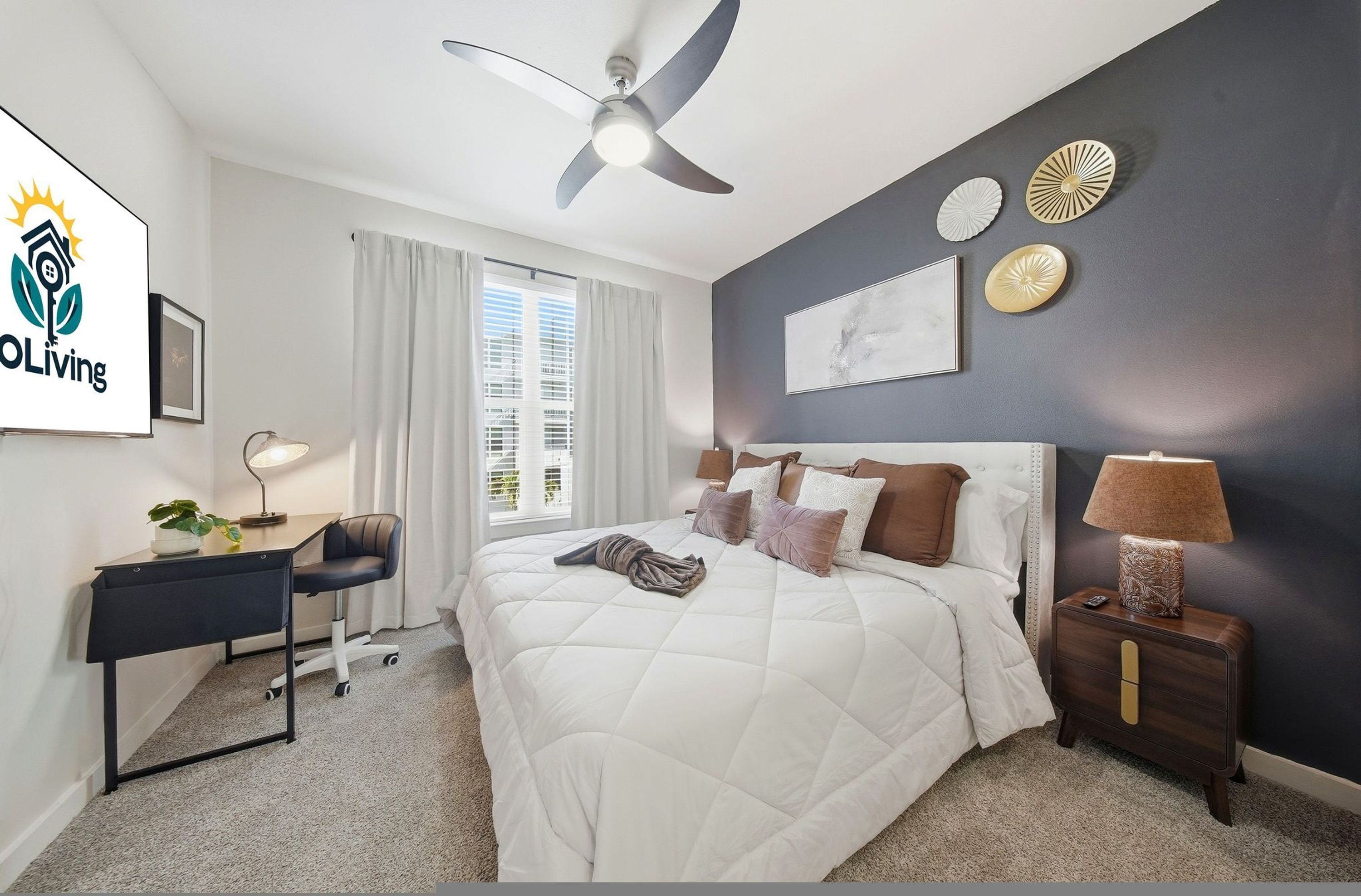 A bedroom with a bed, desk, chair, nightstand, lamp, ceiling fan, and artwork on the wall.