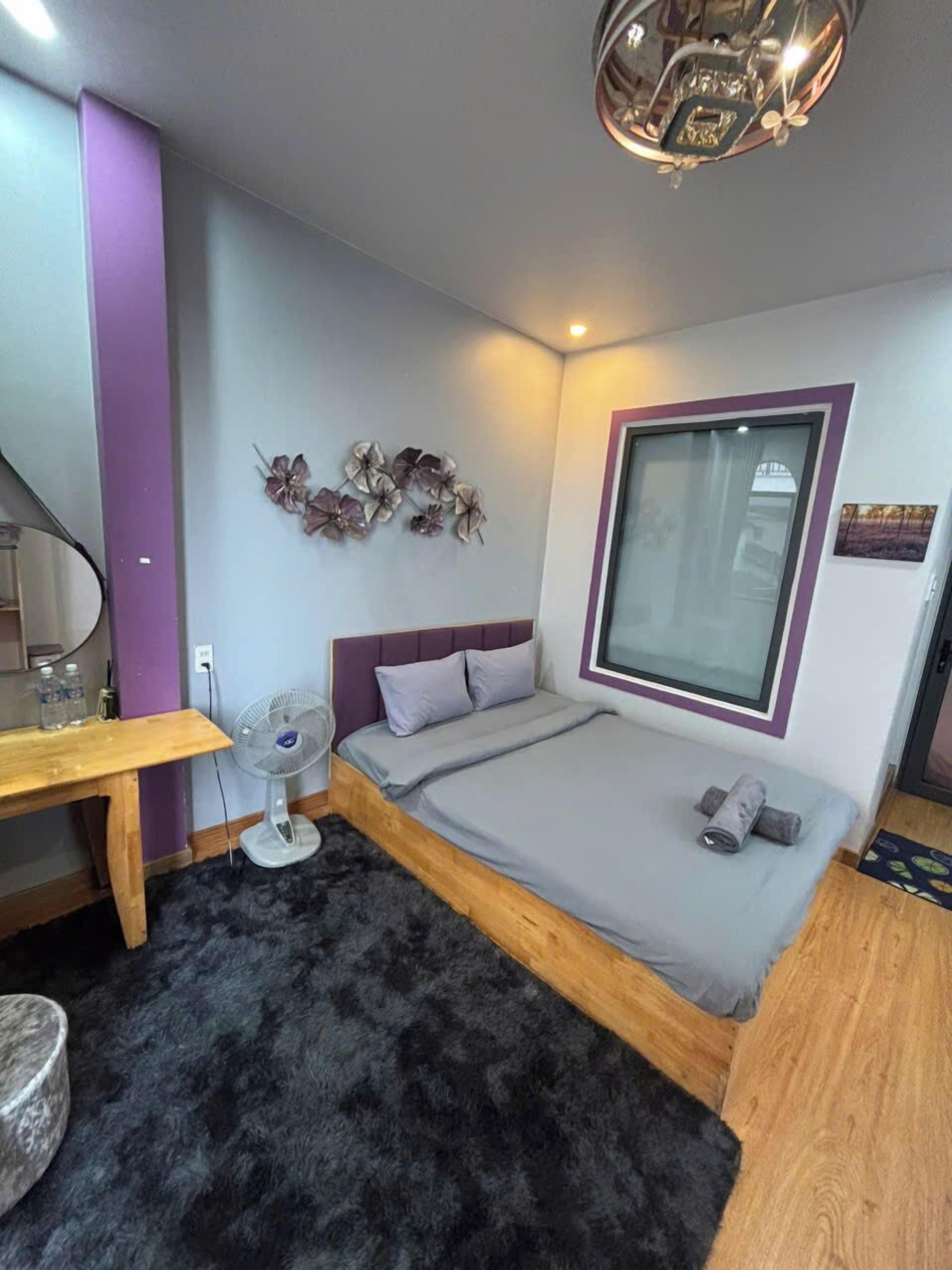 A bedroom with a bed, a desk, a fan, and a window.