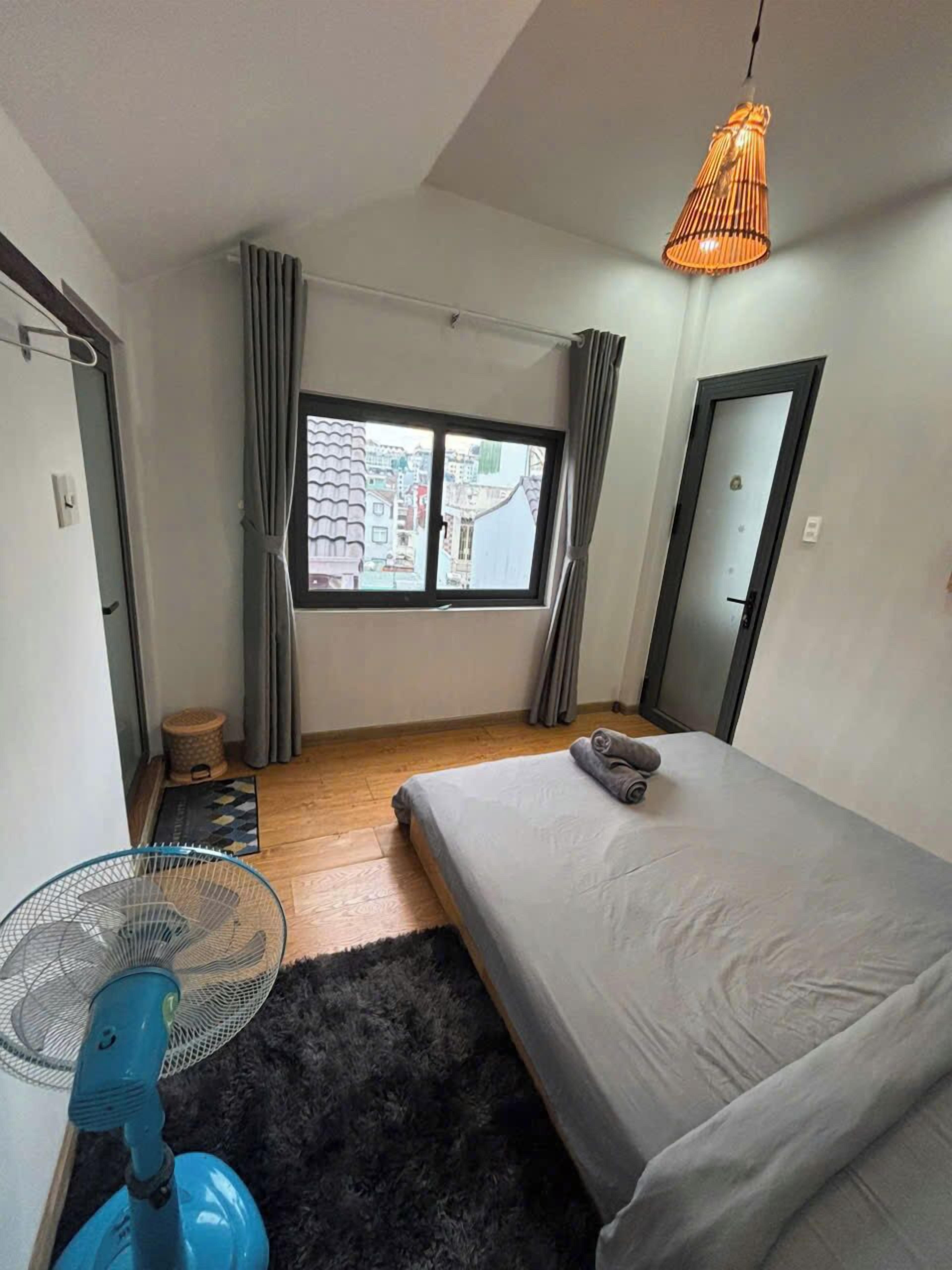 A bedroom with a bed, a window, a fan, and a view of buildings outside.