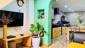 A modern living room with a wooden desk, a large potted plant, a flat-screen TV, and a kitchen area visible through an archway. - Home Nha Gao Da Lat (Xuan Huong - Da Lat)
