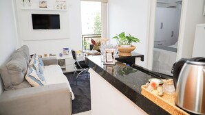 Een moderne woonkamer met een bank, een salontafel, een aan de muur bevestigde televisie, en een keukenhoek met een aanrecht en waterkoker.