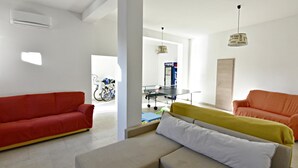 En stue med en rød sofa, en beige sofa og et gult pled. Man kan se et ping pong bord i baggrunden.