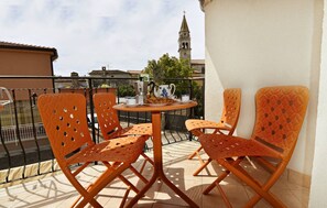 A balcony with orange chairs and a table, a clock, and a view of a tower. - Lovely Poolside Apartment in Tar (Tar-Vabriga)