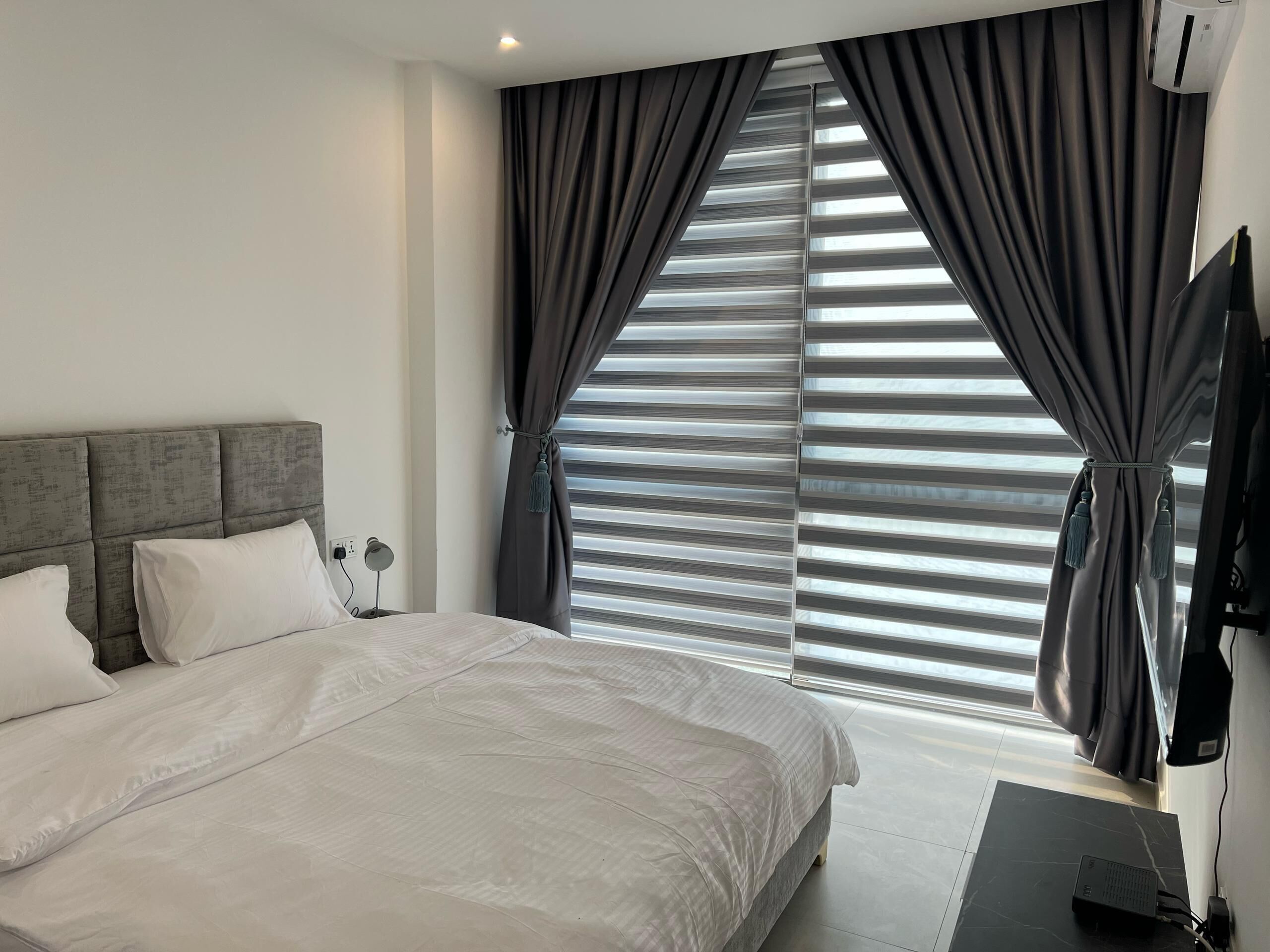 Un dormitorio moderno con una cama grande, cabecera, mesita de noche, televisor montado en la pared y ventana con persianas.