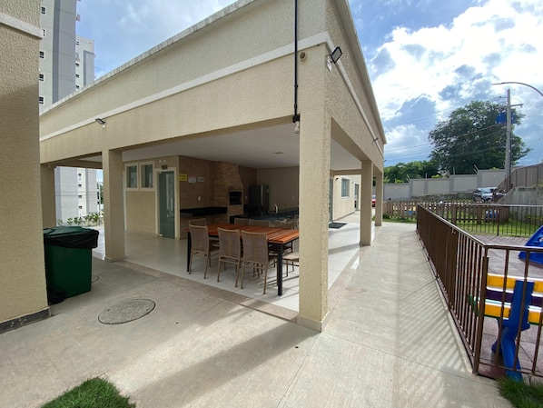 A covered outdoor patio with a dining area, a brick oven, and a view of a residential building. - Cabo Branco Beach. Two-Bedroom Apartment. Up to 7 people. (João Pessoa)