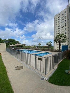 A swimming pool with a metal fence, a grassy area, and a tall building in the background. - Cabo Branco Beach. Two-Bedroom Apartment. Up to 7 people. (João Pessoa)