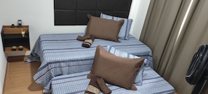 A double bed with striped bedding and two pillows, a bedside table with a remote control, a small shelf with decorative items, and a standing fan. - Cabo Branco Beach. Two-Bedroom Apartment. Up to 7 people. (João Pessoa)