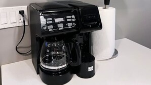 Une machine à café est posée sur un comptoir blanc, au-dessus duquel se trouve une étagère blanche contenant une plante, deux verres à vin et un rouleau d’essuie-tout.