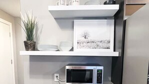 Une cuisine de taille réduite, équipée d’un four à micro-ondes, d’une petite poubelle, d’une plante en pot et d’un tableau encadré posé sur une étagère.