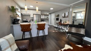 A modern kitchen with white cabinets, a central island, and bar stools. There is a dining area with a black sofa and a cowhide rug. - Dreamy Escape | Cozy & Stylish Stay for Two (London)