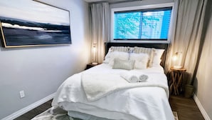 Un lit bien fait, avec une couette blanche et des oreillers, un tapis à motifs, une œuvre d’art accrochée au mur et une fenêtre donnant sur des arbres.